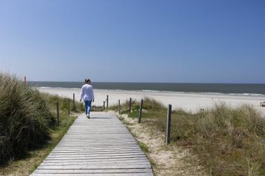 Frau spaziert entlang eines Holzbohlenwegs zum Strand auf Spiekeroog bei blauem Himmel